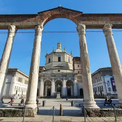 Le imponenti colonne di San Lorenzo incorniciano la piazza con la Basilica di San Lorenzo Maggiore sullo sfondo, sotto un cielo azzurro.