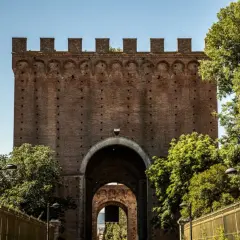 Imponente porta romana in mattoni con archi sovrapposti, circondata da alberi verdi e una recinzione dorata.