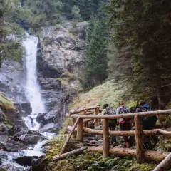 Escursionisti su un sentiero con staccionata in legno ammirano le maestose cascate di Saent, immerse nel verde di un bosco alpino.