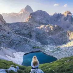 Donna con lunghi capelli biondi ammira un lago alpino nel Parco Nazionale del Tricorno, circondato da maestose vette rocciose illuminate dal sole.