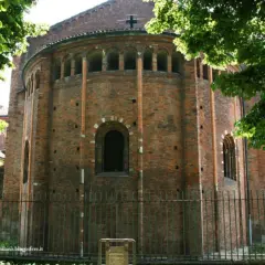 Abside in mattoni di San Nazaro in Brolo, con archetti pensili e una finestra ad arco. Alberi verdi incorniciano la scena.
