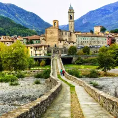 Ponte in pietra attraversa il letto del fiume nel **Val Trebbia**, conducendo a un borgo con campanile e case in pietra sotto montagne verdi.