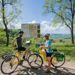 Coppia in bicicletta sulla ciclabile Parenzana, con mappa del percorso e paesaggio collinare sullo sfondo.
