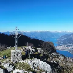 Due escursionisti celebrano la vetta dei Corni di Canzo con una croce e un panorama mozzafiato sul lago.