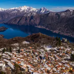 Paesaggio invernale nel Val d'Intelvi con case innevate, lago blu e montagne maestose sullo sfondo.