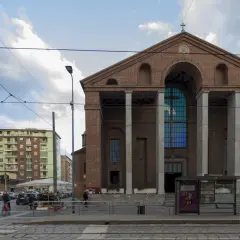 La facciata in mattoni rossi di una chiesa a Milano, con un grande rosone blu e colonne imponenti. Sullo sfondo, palazzi residenziali e tram.