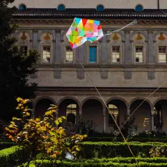 Coloratissimi aquiloni volano nel chiostro dell'Incoronata a Milano, tra siepi curate e un antico porticato.
