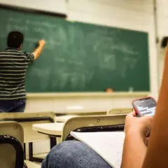 Studente distratto al cellulare mentre il professore scrive alla lavagna, forse un nuovo concorso scuola in arrivo.