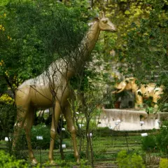 Una giraffa dorata emerge tra la vegetazione lussureggiante dell'orto botanico di Brera, con un elefante dorato sullo sfondo.