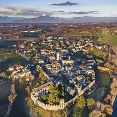 Il borgo medievale di Castellaro Lagusello, con le sue mura e torri, si affaccia su un lago sereno, circondato da colline verdi e montagne all'orizzonte.