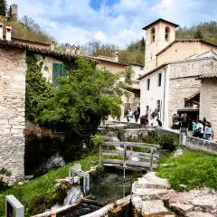 Un borgo in Umbria con cascate che scendono tra case di pietra e un ponte di legno.