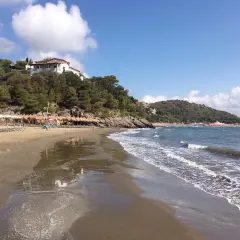 Spiaggia di **Porto d'Ercole** con ombrelloni, lettini e mare calmo sotto un cielo azzurro con nuvole bianche.