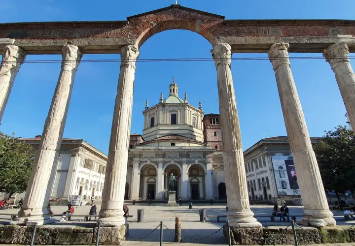 Le imponenti colonne di San Lorenzo incorniciano la piazza con la Basilica di San Lorenzo Maggiore sullo sfondo, sotto un cielo azzurro.