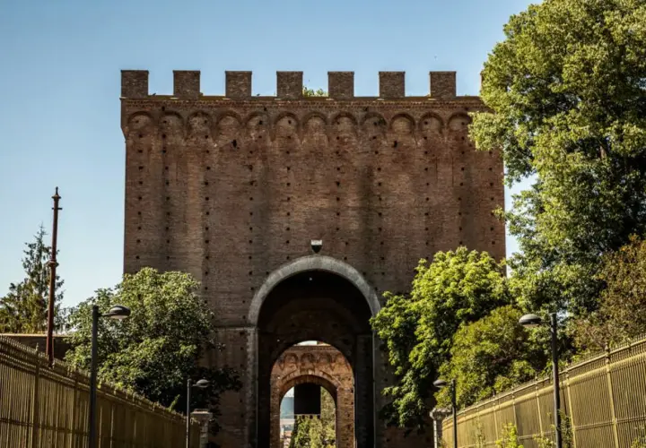 Imponente porta romana in mattoni con archi sovrapposti, circondata da alberi verdi e una recinzione dorata.