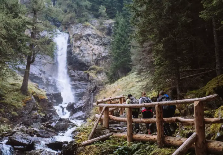 Escursionisti su un sentiero con staccionata in legno ammirano le maestose cascate di Saent, immerse nel verde di un bosco alpino.