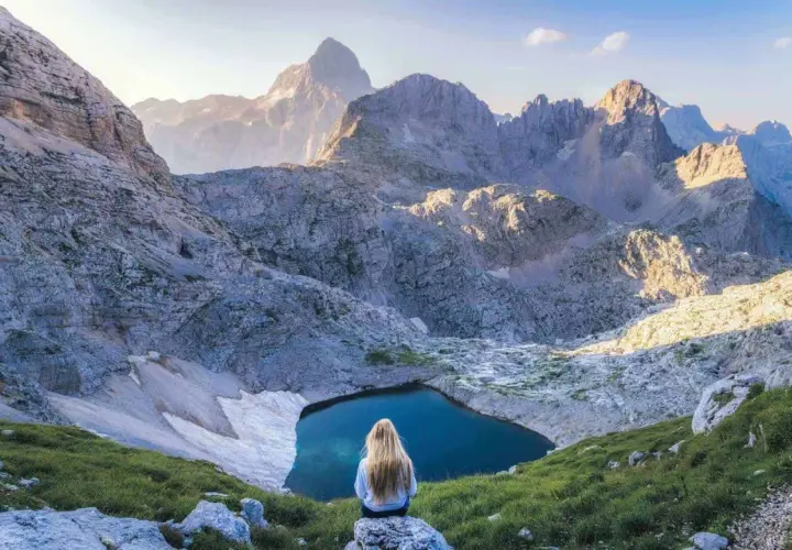 Donna con lunghi capelli biondi ammira un lago alpino nel Parco Nazionale del Tricorno, circondato da maestose vette rocciose illuminate dal sole.