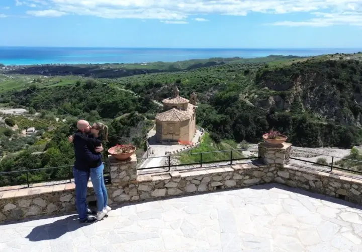 Coppia si bacia con vista panoramica su Badolato, Calabria, con il mare all'orizzonte e una chiesa antica.
