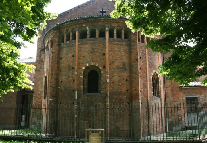 Abside in mattoni di San Nazaro in Brolo, con archetti pensili e una finestra ad arco. Alberi verdi incorniciano la scena.