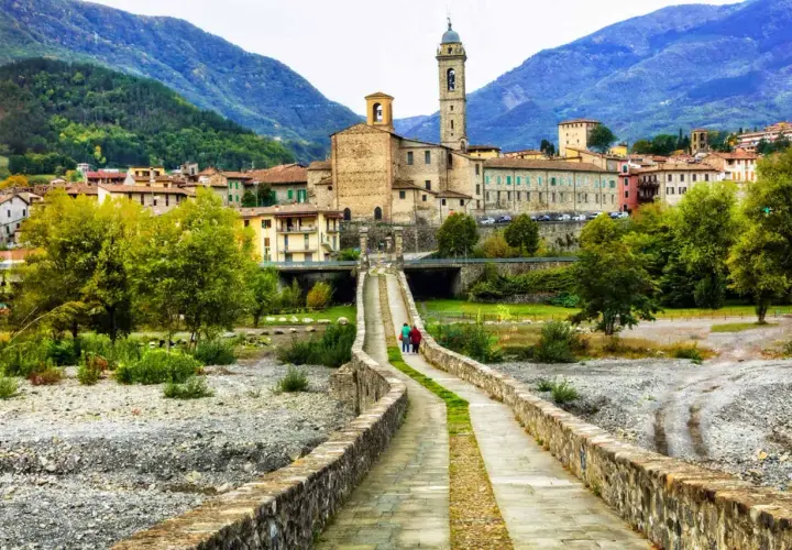 Ponte in pietra attraversa il letto del fiume nel **Val Trebbia**, conducendo a un borgo con campanile e case in pietra sotto montagne verdi.