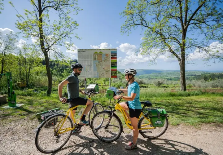 Coppia in bicicletta sulla ciclabile Parenzana, con mappa del percorso e paesaggio collinare sullo sfondo.