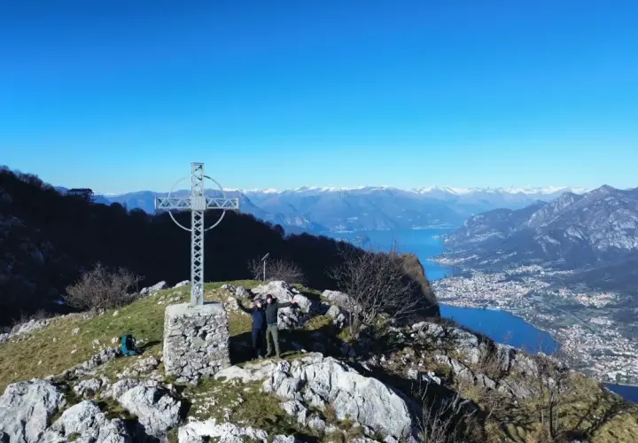 Due escursionisti celebrano la vetta dei Corni di Canzo con una croce e un panorama mozzafiato sul lago.