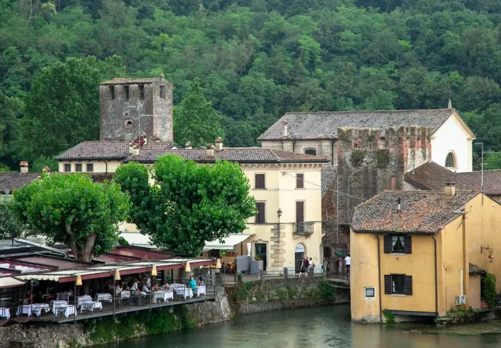 Un pittoresco borghetto sul Mincio con case storiche, torri medievali e un ristorante con tavoli all'aperto affacciato sul fiume.