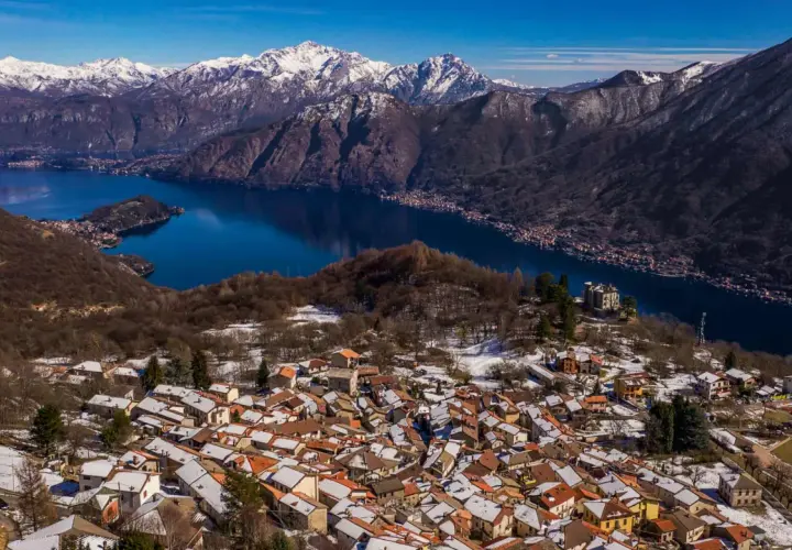 Paesaggio invernale nel Val d'Intelvi con case innevate, lago blu e montagne maestose sullo sfondo.