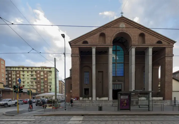 La facciata in mattoni rossi di una chiesa a Milano, con un grande rosone blu e colonne imponenti. Sullo sfondo, palazzi residenziali e tram.