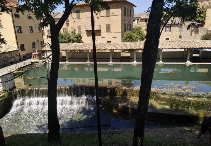 Cascata e portico storico a Bevagna, Umbria. L'acqua scende su gradoni di pietra, riflettendo gli edifici antichi.