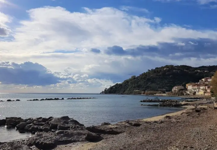 Costa di Pollica con case colorate arroccate sulla collina e mare calmo sotto un cielo nuvoloso.