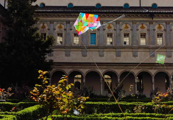 Coloratissimi aquiloni volano nel chiostro dell'Incoronata a Milano, tra siepi curate e un antico porticato.