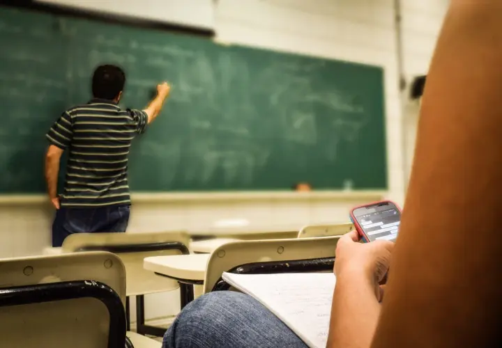 Studente distratto al cellulare mentre il professore scrive alla lavagna, forse un nuovo concorso scuola in arrivo.