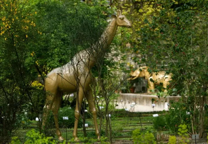 Una giraffa dorata emerge tra la vegetazione lussureggiante dell'orto botanico di Brera, con un elefante dorato sullo sfondo.