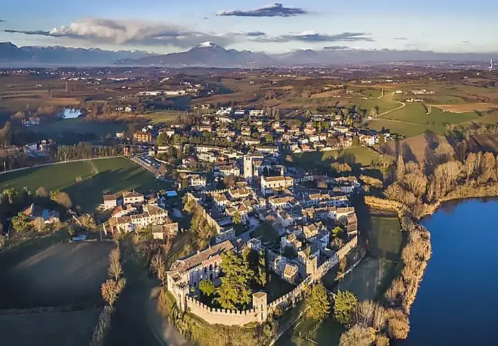 Il borgo medievale di Castellaro Lagusello, con le sue mura e torri, si affaccia su un lago sereno, circondato da colline verdi e montagne all'orizzonte.