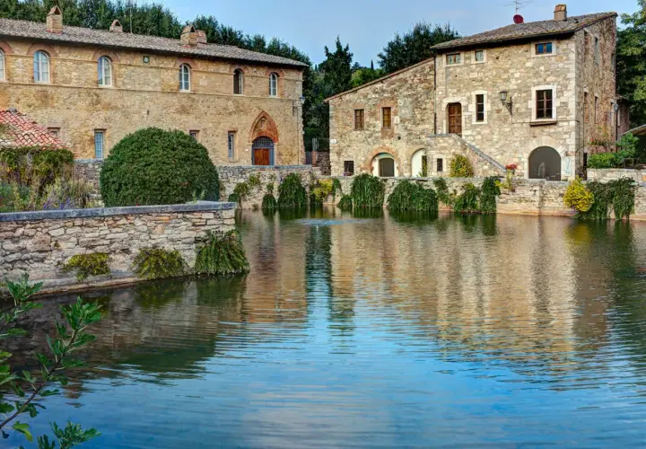 Antiche terme libere a San Gimignano, con edifici in pietra che si specchiano in un laghetto tranquillo.
