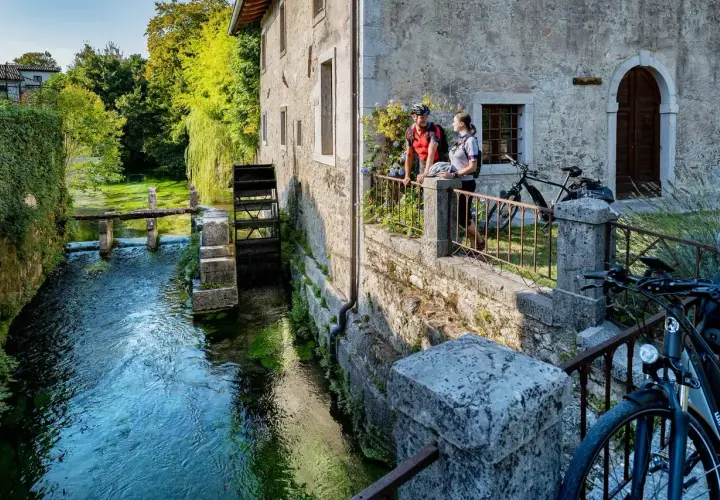 Coppia in bicicletta ammira un antico mulino ad acqua, uno dei tanti borghi friulani da visitare, immersi nel verde e lungo un ruscello cristallino.