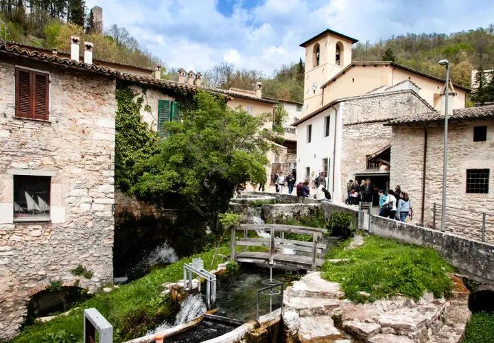 Un borgo in Umbria con cascate che scendono tra case di pietra e un ponte di legno.