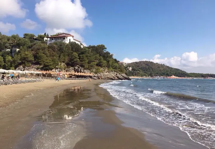 Spiaggia di **Porto d'Ercole** con ombrelloni, lettini e mare calmo sotto un cielo azzurro con nuvole bianche.