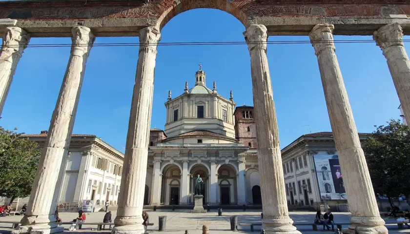 Le imponenti colonne di San Lorenzo incorniciano la piazza con la Basilica di San Lorenzo Maggiore sullo sfondo, sotto un cielo azzurro.