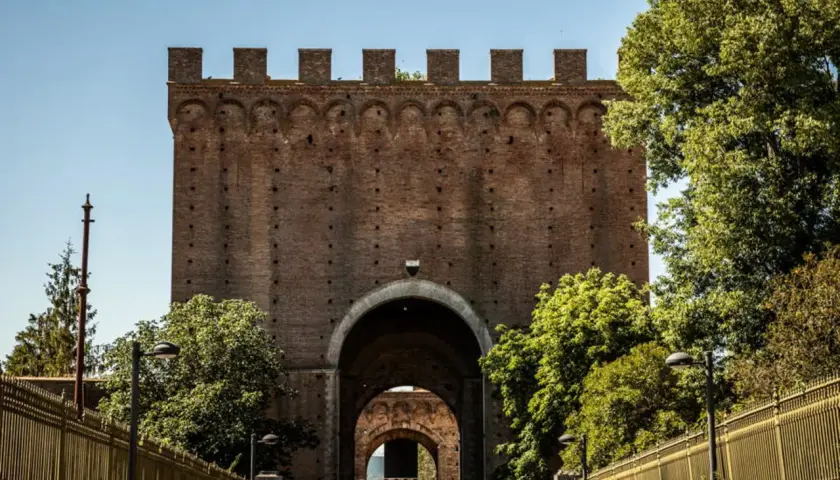 Imponente porta romana in mattoni con archi sovrapposti, circondata da alberi verdi e una recinzione dorata.