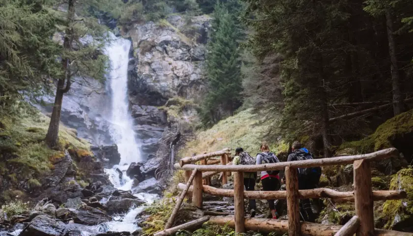 Escursionisti su un sentiero con staccionata in legno ammirano le maestose cascate di Saent, immerse nel verde di un bosco alpino.