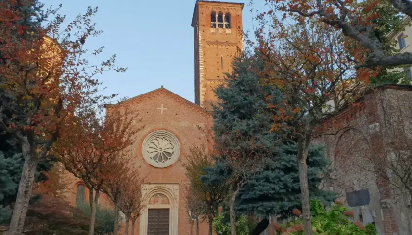San Celso a Milano, con il suo campanile in mattoni rossi e la facciata ornata da un rosone, circondato da alberi dalle foglie autunnali.