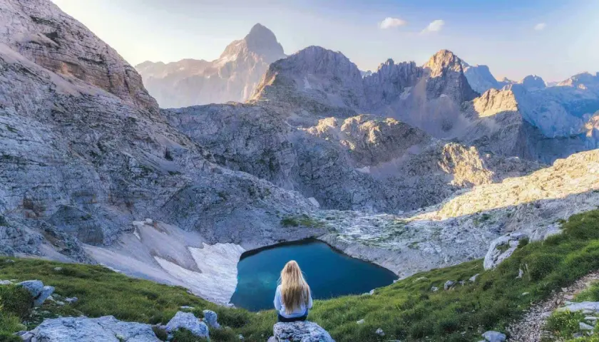 Donna con lunghi capelli biondi ammira un lago alpino nel Parco Nazionale del Tricorno, circondato da maestose vette rocciose illuminate dal sole.