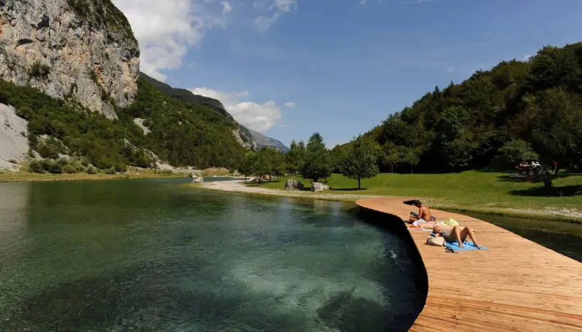 Persone si rilassano sul pontile in legno del lago di Nembia, circondato da montagne verdi e acqua cristallina.