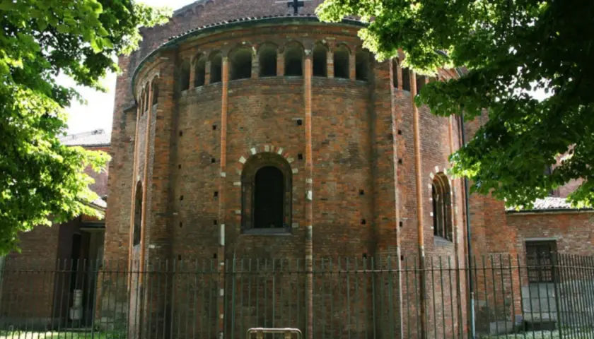 Abside in mattoni di San Nazaro in Brolo, con archetti pensili e una finestra ad arco. Alberi verdi incorniciano la scena.