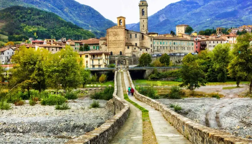 Ponte in pietra attraversa il letto del fiume nel **Val Trebbia**, conducendo a un borgo con campanile e case in pietra sotto montagne verdi.