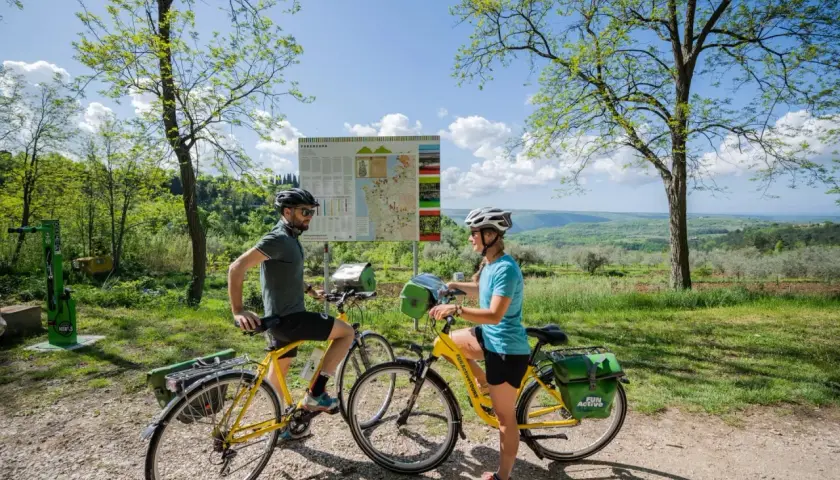 Coppia in bicicletta sulla ciclabile Parenzana, con mappa del percorso e paesaggio collinare sullo sfondo.