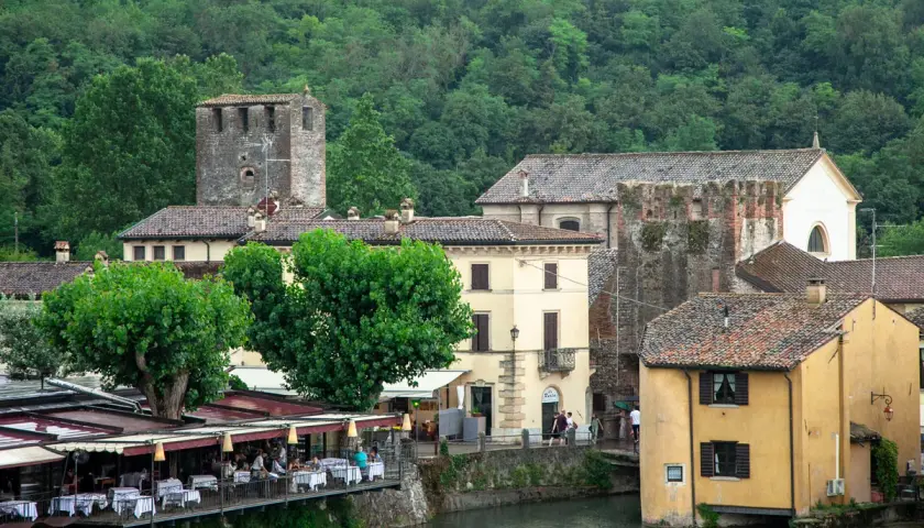 Un pittoresco borghetto sul Mincio con case storiche, torri medievali e un ristorante con tavoli all'aperto affacciato sul fiume.