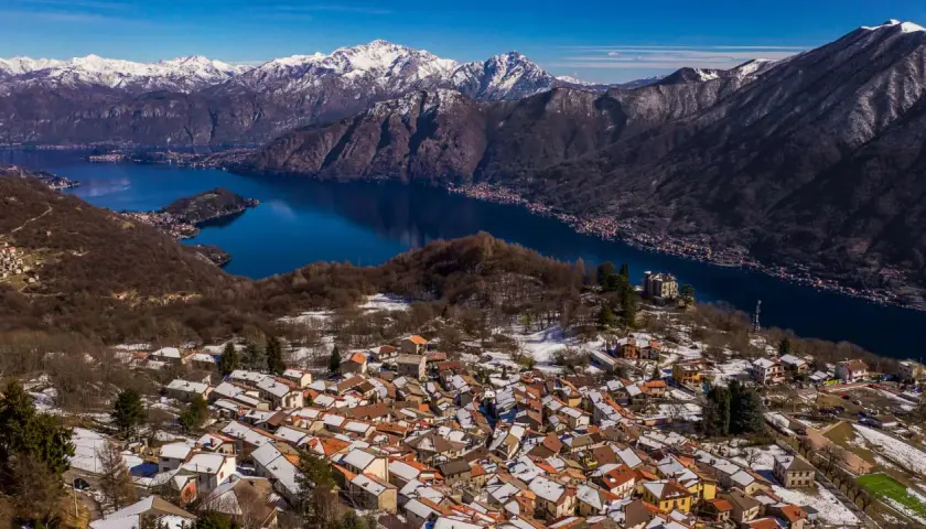 Paesaggio invernale nel Val d'Intelvi con case innevate, lago blu e montagne maestose sullo sfondo.