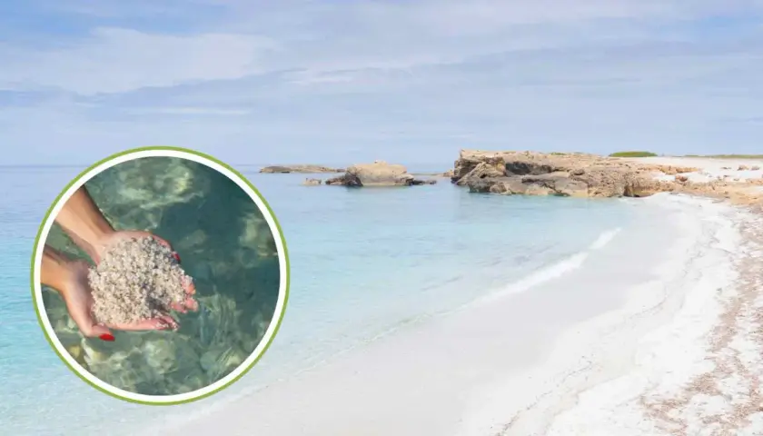 Mani raccolgono sabbia bianca e fine su una spiaggia di vetro in Sardegna, con acque cristalline e rocce sullo sfondo.